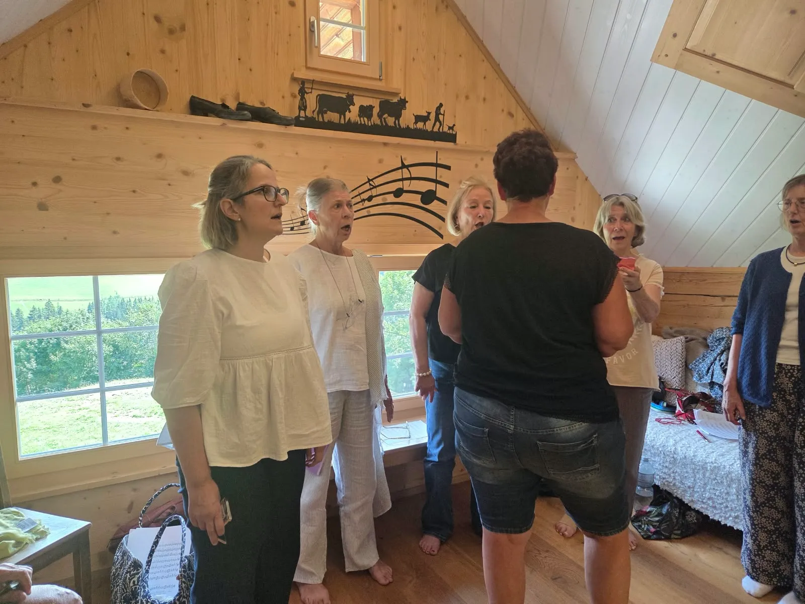 Des stagiaires chantent dans une pièce en bois sous les combles d'un chalet, une grande fenêtre ouverte sur la prairie.