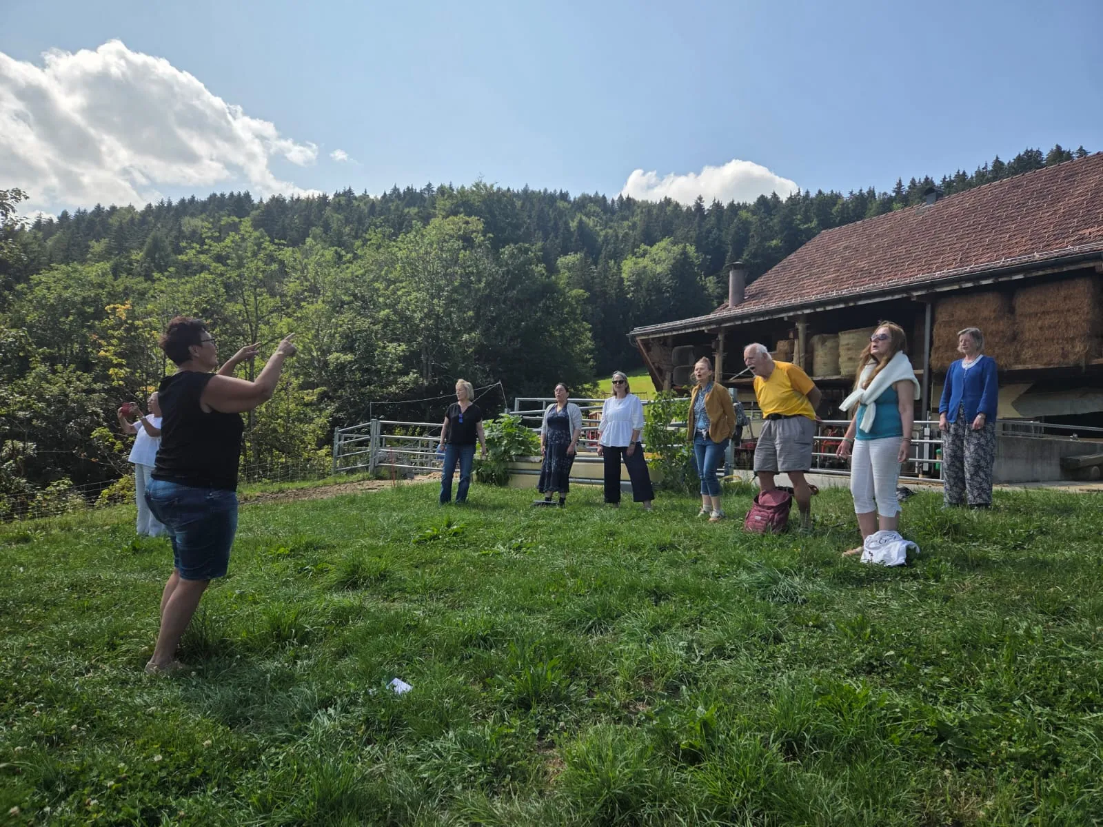 Une répétition en plein air, dans l'herbe devant une ferme, la cheffe de chœur face au groupe.